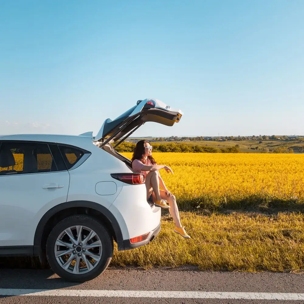Lächelnde huebsche Frau, die bei Sonnenuntergang im Kofferraum des SUV sitzt