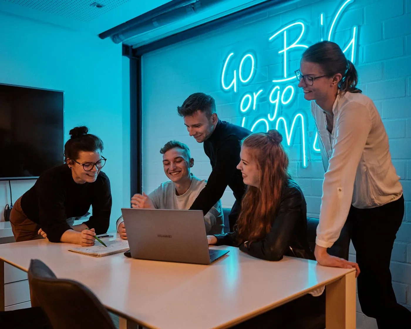Suchhelden-Team bespricht ein Projekt am Laptop vor dem Neonschriftzug „Go big or go home“ in…