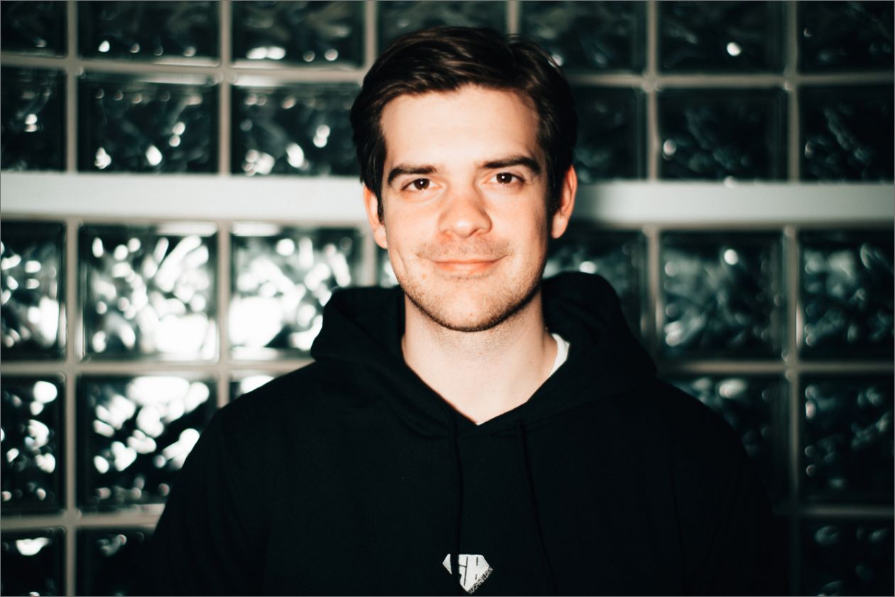 Steven Wart in schwarzem Hoodie lächelt vor einer Wand aus Glasbausteinen.