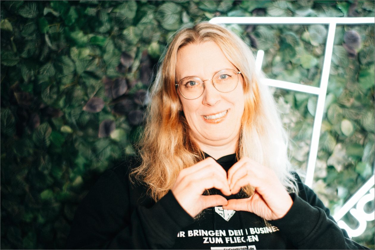 Nadine Sogl formt Herz vor Blatt-Hintergrund, trägt Brille und lächelt freundlich.