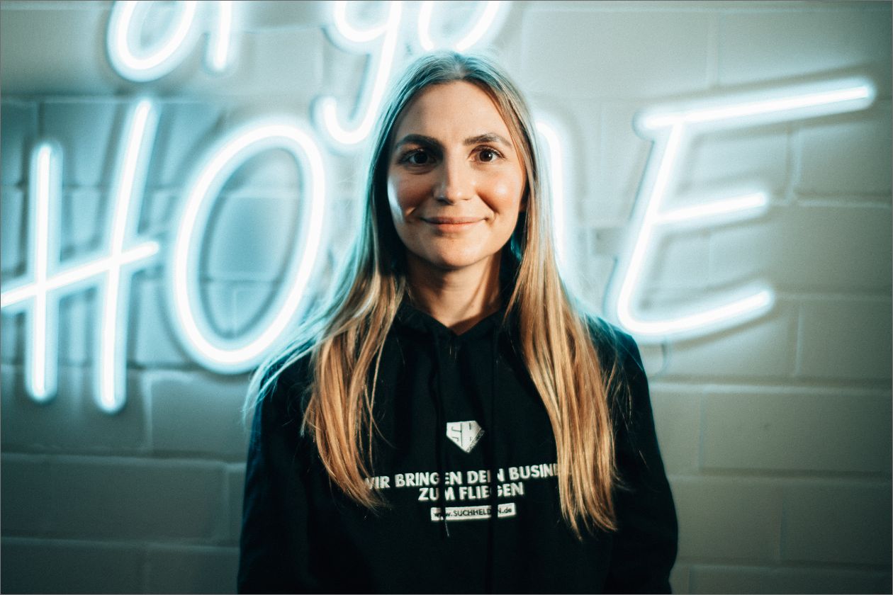 Sandra Nein steht vor einer blau beleuchteten Wand, trägt schwarzen Hoodie.