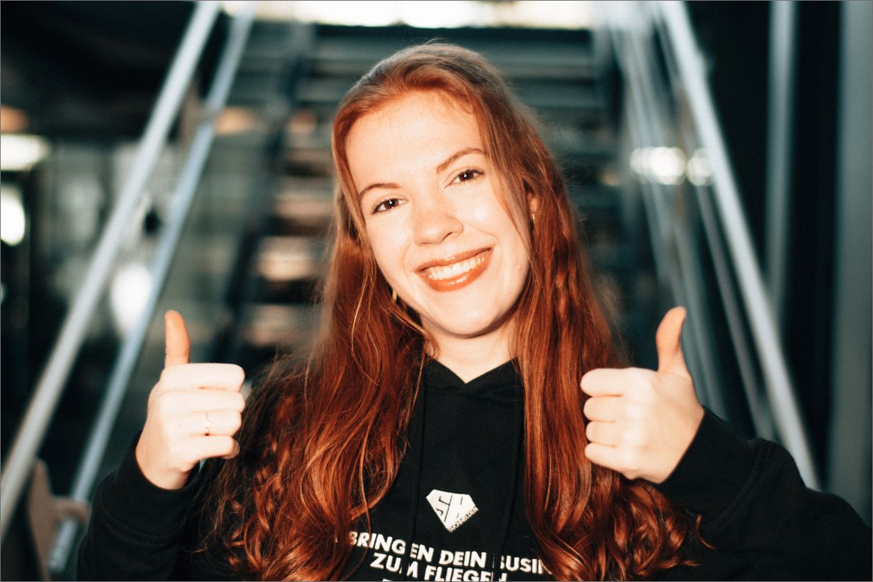Jill Weyers zeigt Daumen hoch, steht vor Treppe, trägt schwarzen Hoodie.