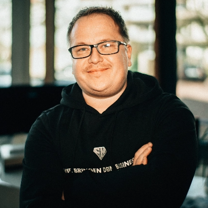 Andre Thyhatmer mit schwarzem Hoodie posiert lächelnd vor einer Fensterwand.