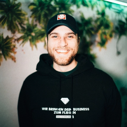 Malte Krekeler lächelt und trägt Kapuzenpullover vor einem tropischem Hintergrund.